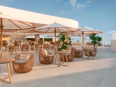 Terrazza aperta con sedie in rattan, tavoli in legno e metallo e grandi ombrelloni sotto un cielo sereno.