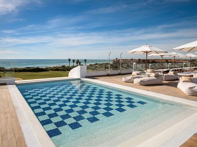 Moderner Pool mit blau-weißem Kachelmuster und Liegen mit Sonnenschirmen, Meer im Hintergrund.
