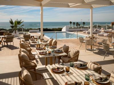 Terrasse eines Hotels mit Tischen und Stühlen, Blick auf Pool und Meer im Hintergrund.