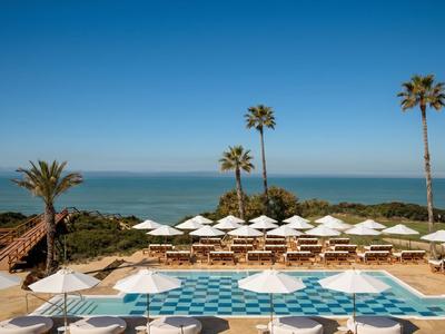 Luxuriöser Pool mit weißen Sonnenschirmen und Palmen vor Meerblick an einem sonnigen Tag.