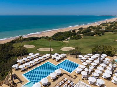 Luxuriöser Hotelpool mit weißen Sonnenschirmen am Meer und Golfplatz im Hintergrund.