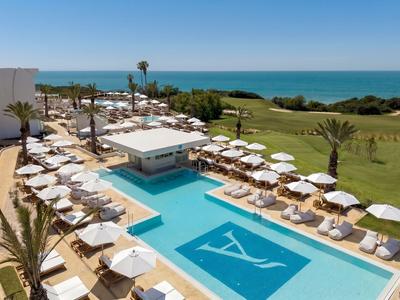 Luxuriöser Poolbereich mit weißen Liegestühlen, Sonnenschirmen und Blick auf das Meer und Grünflächen.