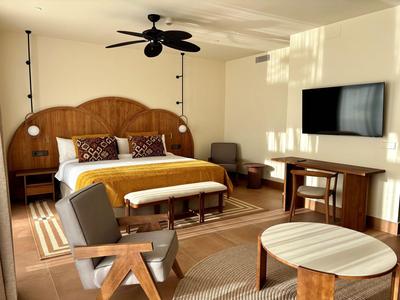 Helles Hotelzimmer mit großem Bett, zwei grauen Sesseln, Deckenventilator und an der Wand montiertem Fernseher.