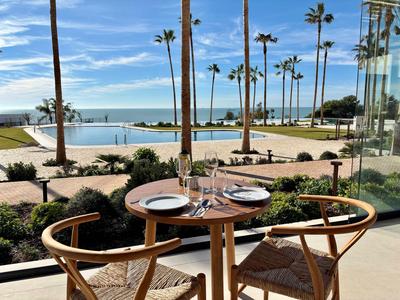 Tisch mit zwei Stühlen auf Terrasse mit Blick auf Pool, Palmen und Meer bei blauem Himmel.