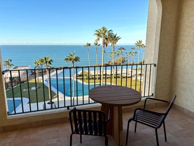 Balkon mit Stühlen, Tisch und Blick auf Pool, Palmen und Meer an einem sonnigen Tag.