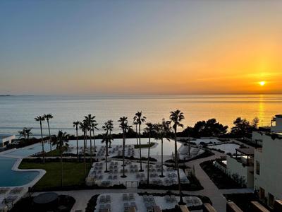 Strandhotel mit Pool, Palmen und Sonnenuntergang über ruhigem Meer.