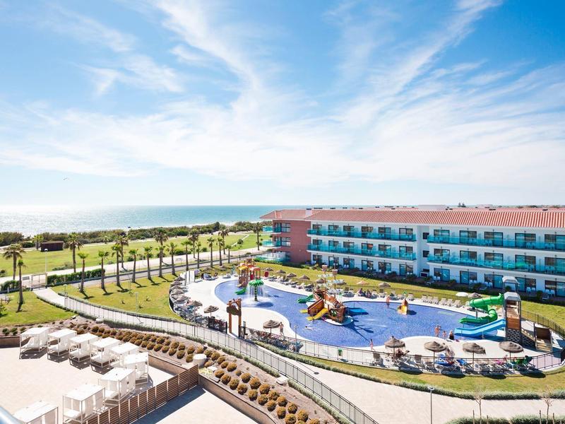 Hotel con piscina all'aperto, scivoli d'acqua, palme e vista sull'oceano in giornata di sole.