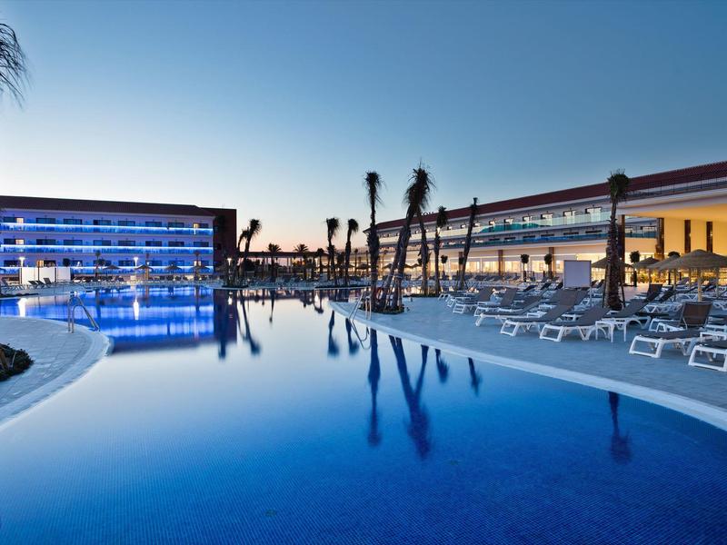 Piscina all'aperto al tramonto con file di lettini e palme.