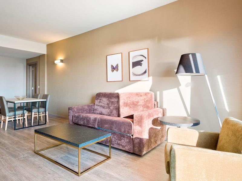 Camera d'hotel luminosa con divano rosa, poltrona beige, tavolo da pranzo in vetro e decorazioni murali moderne.