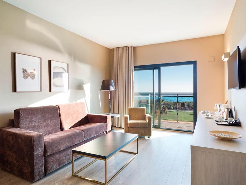 Camera d'hotel luminosa con divano, poltrona, tavolino, TV e vista mare dal balcone.