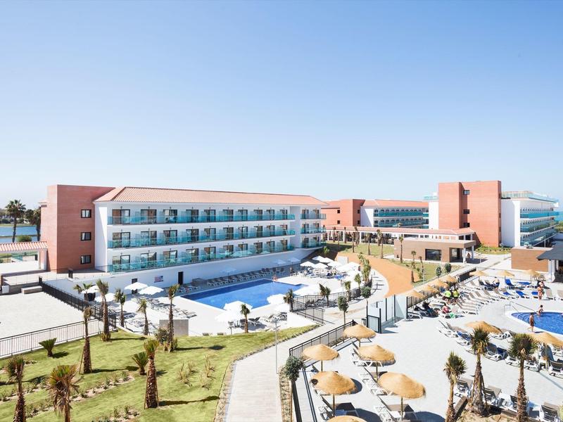 Hotel moderno con piscine, lettini e palme vicino al mare sotto un cielo azzurro chiaro.