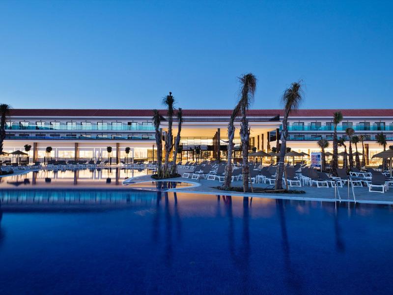 Hotel moderno con piscina illuminata e palme al crepuscolo sotto un cielo terso.
