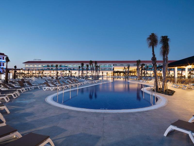 Vista serale di una piscina curva dell'hotel con lettini e palme sotto un cielo blu chiaro.