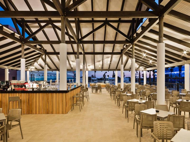 Ampia area pranzo all'aperto con sedie in vimini e bar in legno sotto un grande tetto inclinato al crepuscolo.