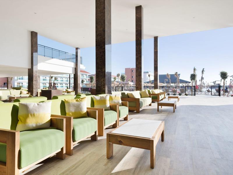 Area lounge all'aperto elegante con poltrone verdi imbottite e tavoli in legno sotto un tetto ombreggiato.