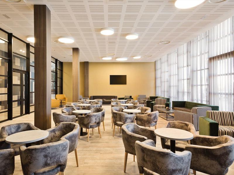 Lounge d'hotel moderno con tavoli rotondi, poltrone imbottite e grandi finestre luminose.