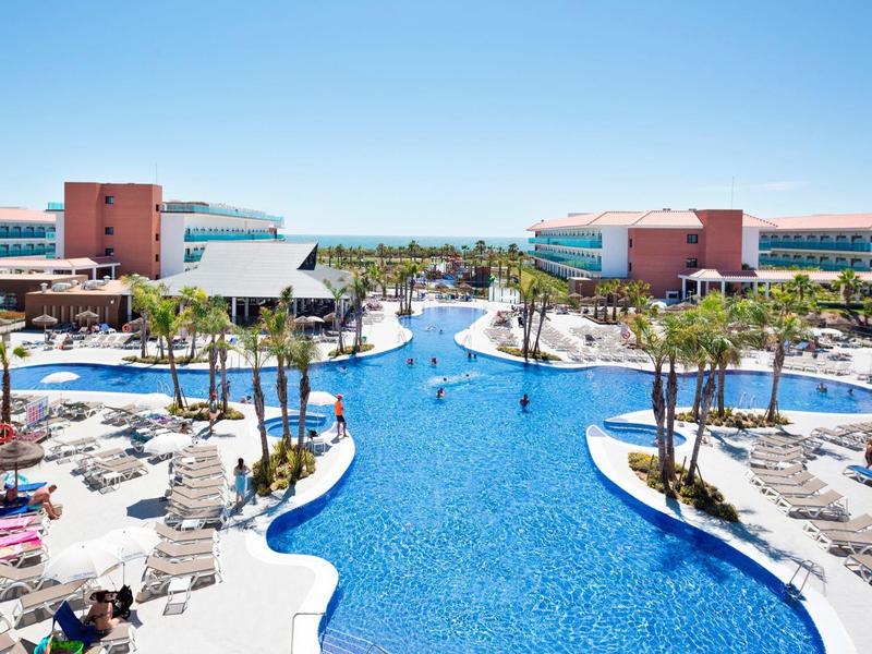 Resort di lusso con grande piscina, lettini, palme e vista mare sotto un cielo sereno.