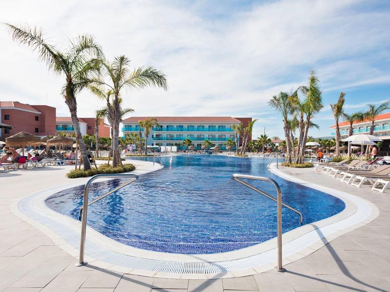 Piscina esterna dell'hotel con palme e lettini sotto un cielo azzurro.