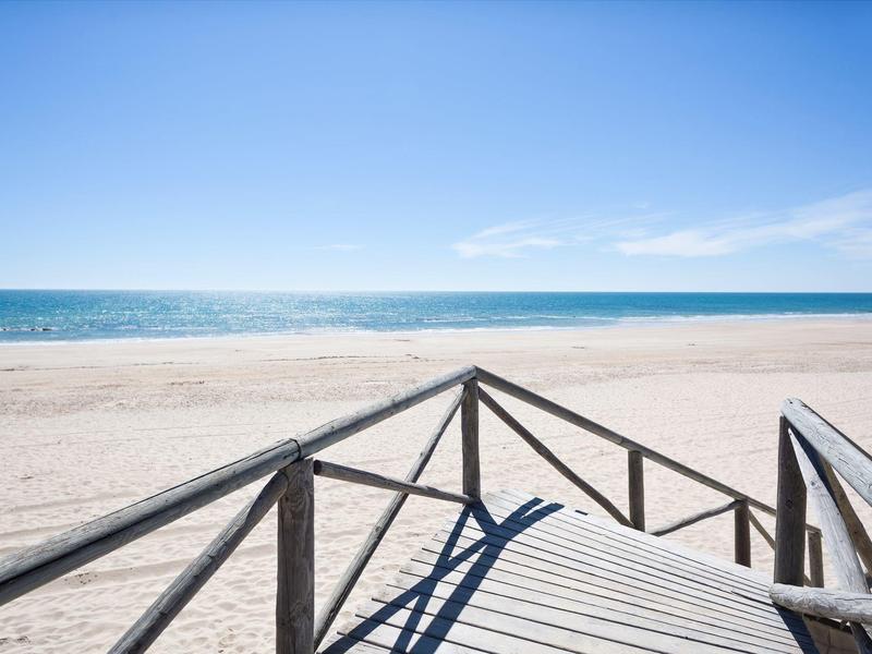 Passerella in legno che conduce a una spiaggia assolata e vuota con cielo azzurro e oceano.