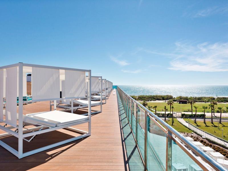 Lettini con baldacchini su una terrazza in legno con vista sul mare e sulle palme.