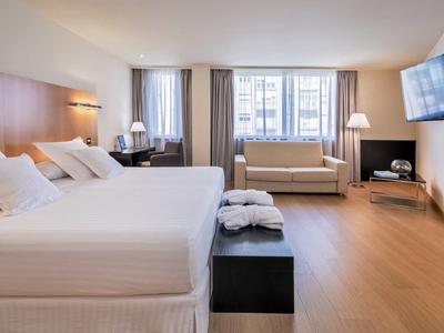 Habitación de hotel luminosa con cama doble, sofá, escritorio y TV en la pared.