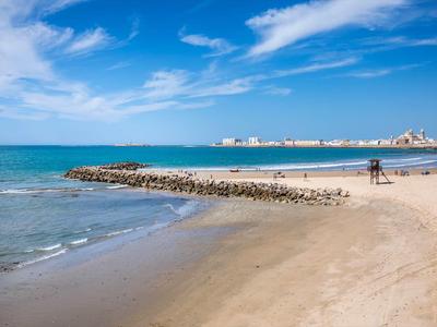 Písčitá pláž s modrou oblohou, vlnami a promenádou v pozadí za slunného dne
