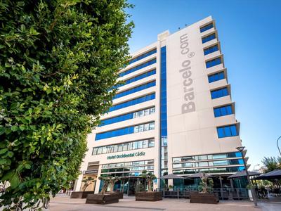 Edificio alto con fachada blanca y ventanas azules bajo un cielo despejado y un árbol frondoso al lado.