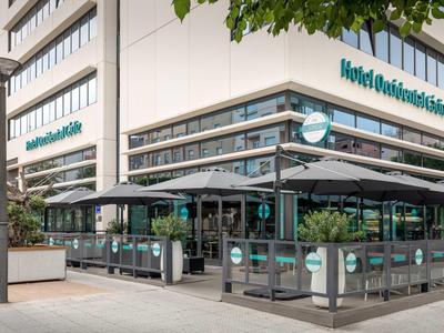 Vstup a terasa moderního městského hotelu s markýzami a skleněnými zábradlími.