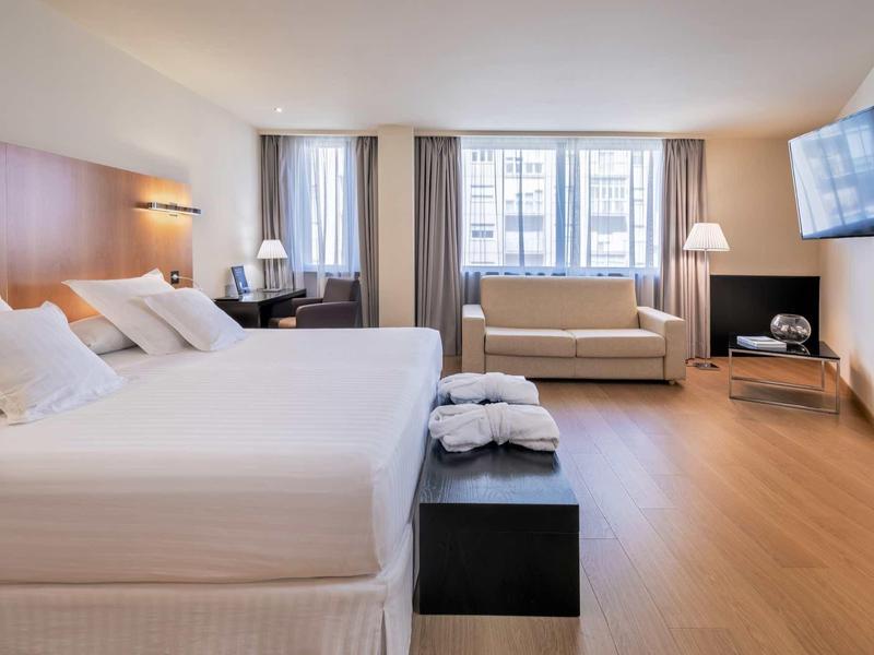 Habitación de hotel luminosa con cama doble, sofá, escritorio y TV en la pared.