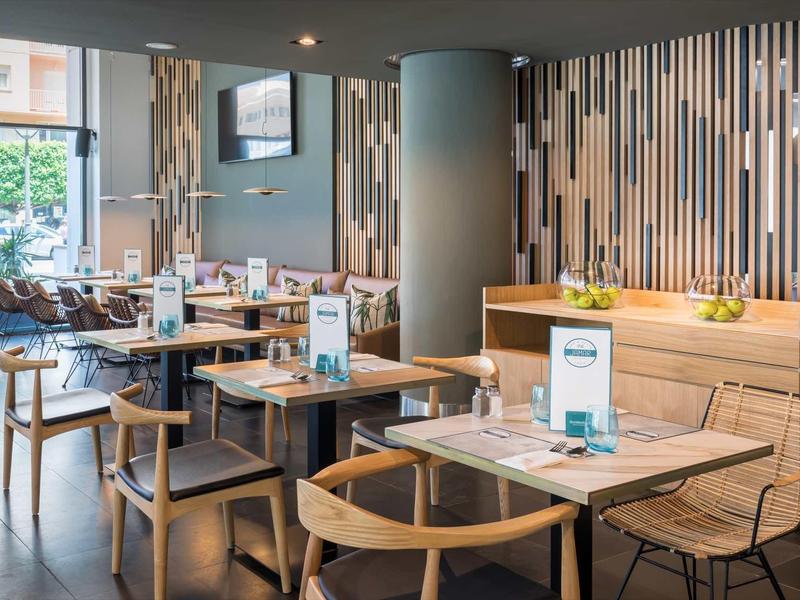 Área de restaurante moderno en hotel con mesas y sillas de madera y elementos decorativos en la pared.