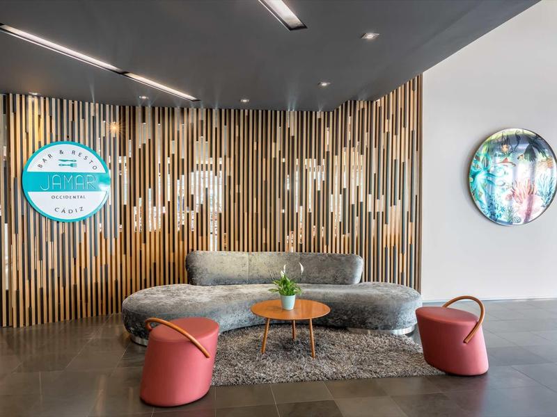 Área lounge moderna con sofá gris, dos pufs rojos, revestimiento de madera y espejo en la pared.