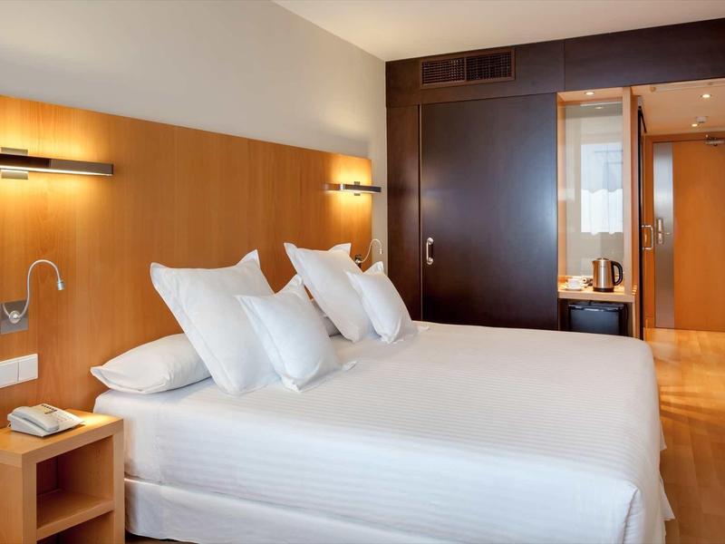 Habitación de hotel moderna con cama doble, almohadas blancas, muebles de madera e iluminación indirecta.