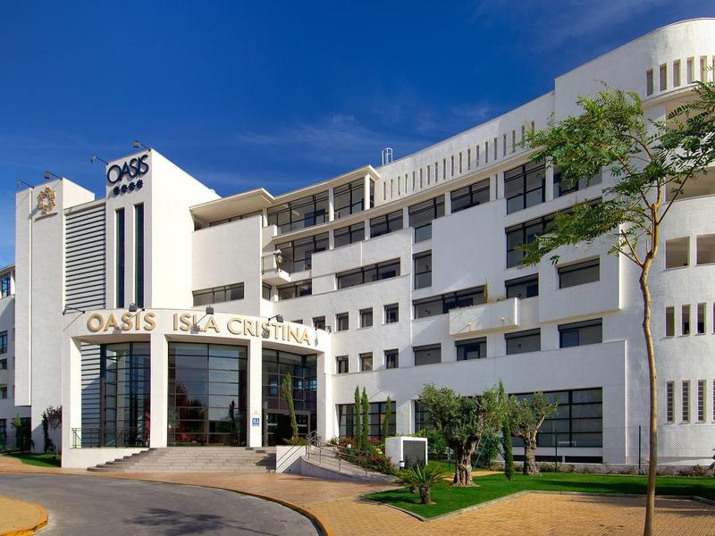 Moderní bílá hotelová budova s zaoblenými balkony a velkým vstupem pod jasnou oblohou.