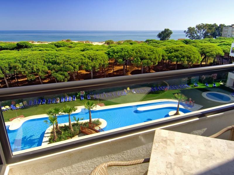 Balkon s výhledem na bazén, zelené stromy a moře v pozadí.