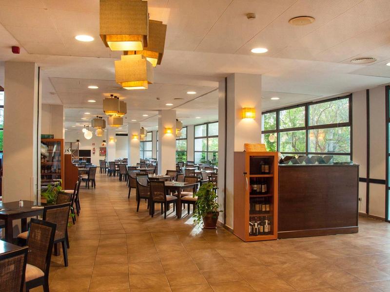 Velká světlá jídelna s dřevěnými židlemi a stoly, moderním osvětlením a velkými okny.