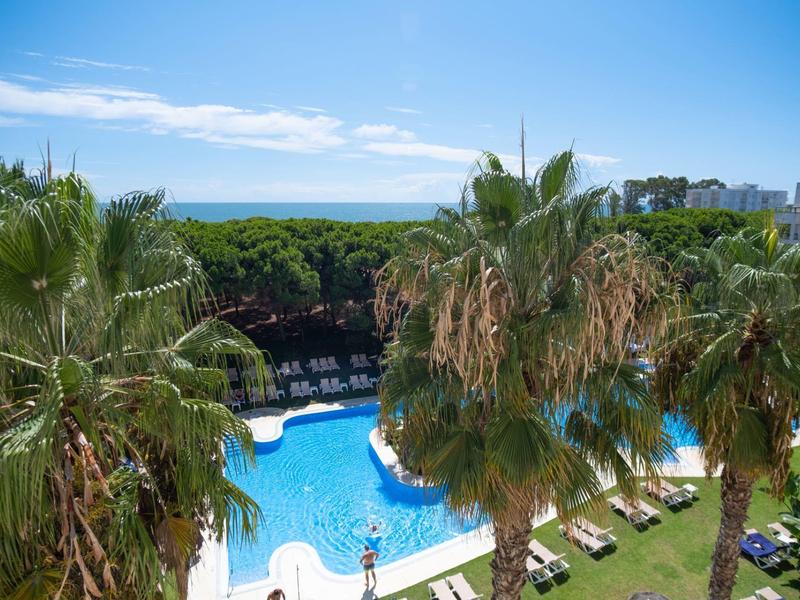 Velký venkovní bazén s lehátky, obklopený palmami s výhledem na pobřeží.