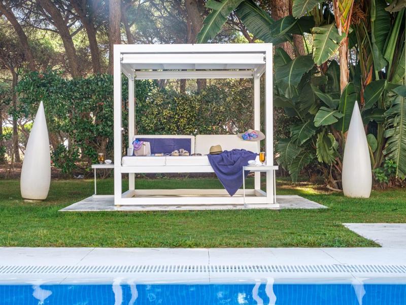 Moderní venkovní posezení s bílým denním lůžkem a modrou dekou vedle bazénu.