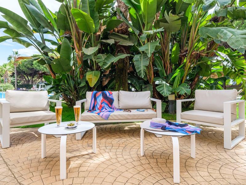 Moderní venkovní posezení s bílým nábytkem a zelenými rostlinami v pozadí.