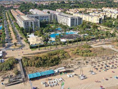 Vista aérea de un hotel con piscina junto a una extensa playa de arena con tumbonas y sombrillas.