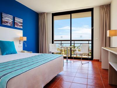 Habitación de hotel luminosa con vista al mar, cama doble, balcón y decoración moderna.