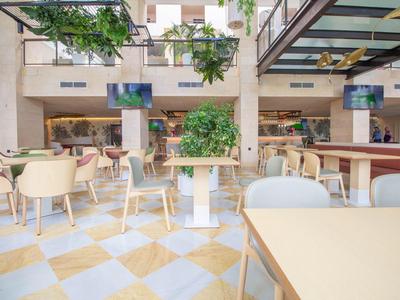 Área interior luminosa y abierta de hotel con mesas blancas, sillas y plantas.