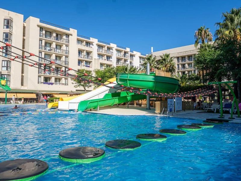 Piscina de hotel con tobogán acuático y piedras flotantes bajo cielo azul despejado.