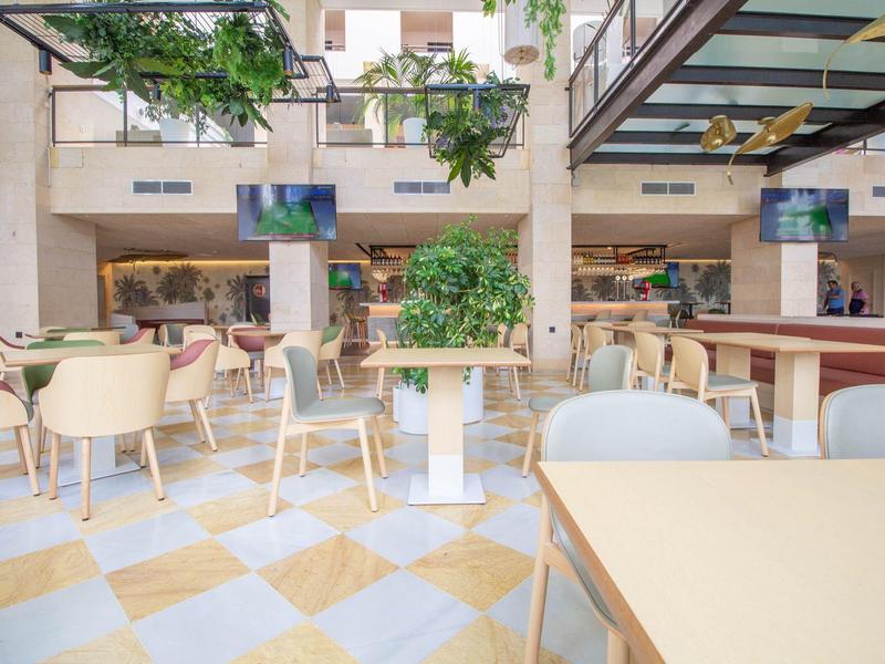 Área interior luminosa y abierta de hotel con mesas blancas, sillas y plantas.