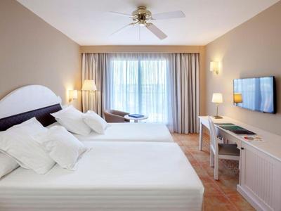 Camera d'hotel luminosa con due grandi letti, ventilatore a soffitto e balcone con tende.