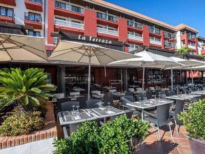 Vista esterna di un hotel con una terrazza piena di tavoli e sedie sotto grandi ombrelloni.