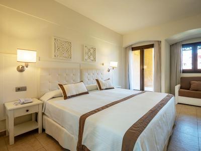 Acogedora habitación doble con paredes claras, dos camas, mesas de noche y sofá junto a la ventana.