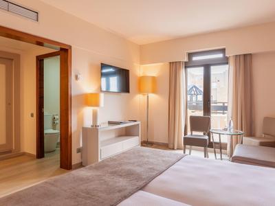 Habitación de hotel luminosa con dos camas, área de estar y balcón con vista al exterior.