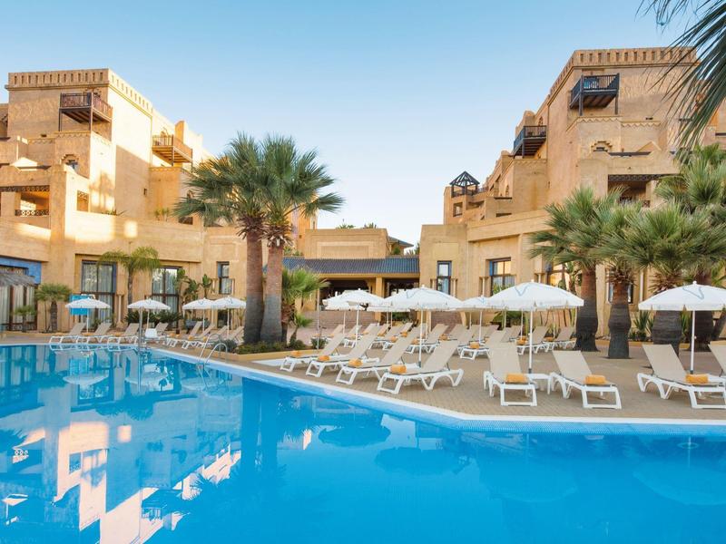 Hotel con piscina azul, tumbonas y palmeras bajo un cielo claro durante el día.