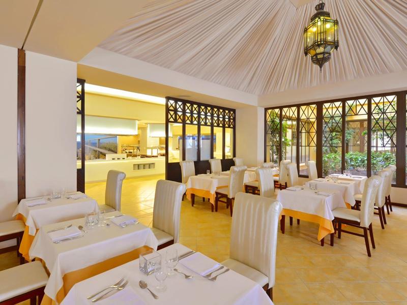 Restaurante luminoso con mesas y sillas blancas, grandes ventanas y iluminación en el techo.