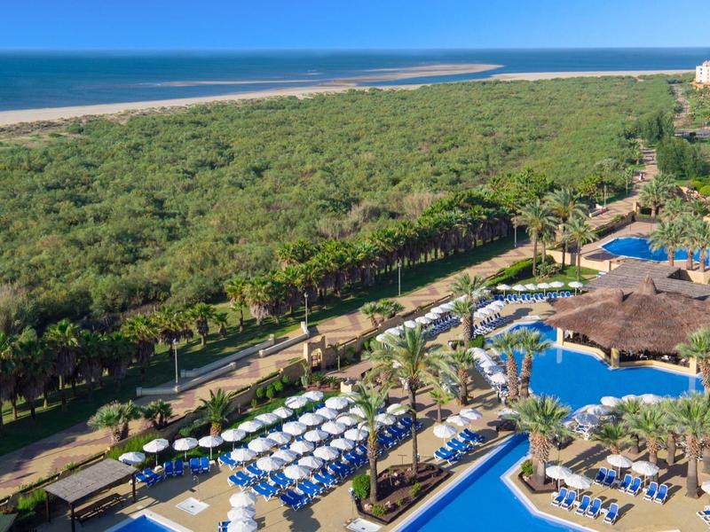 Vista de un hotel con piscina junto a un bosque y playa bajo cielo azul.